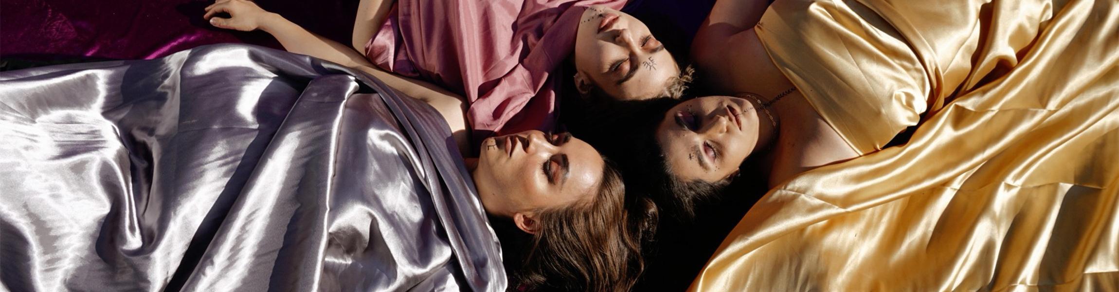 Three Kurdish women lay on their backs, eyes closed and heads all meeting at the center. They are draped in vibrant reflective fabric.
