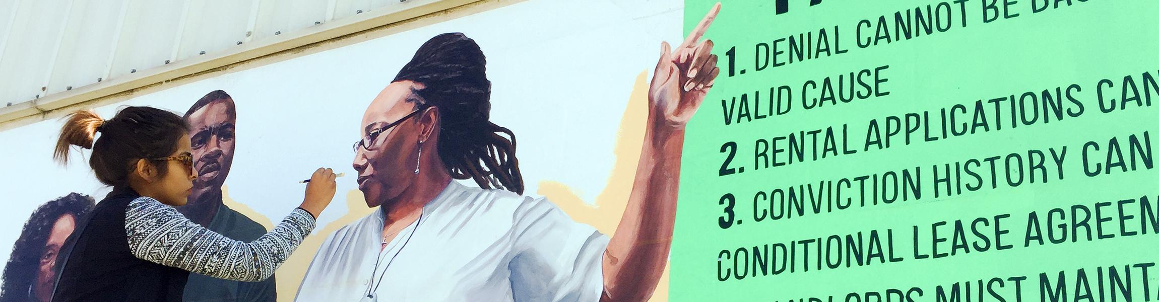 A young woman of color paints a mural. The visible portion of the mural is of a Black woman point to a list of tenants' rights shown in black paint on bright green background.
