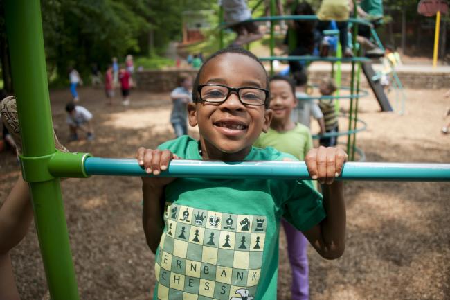 A young Black child wearing glasses smiles a big toothy grin while hanging on a playground bar.
