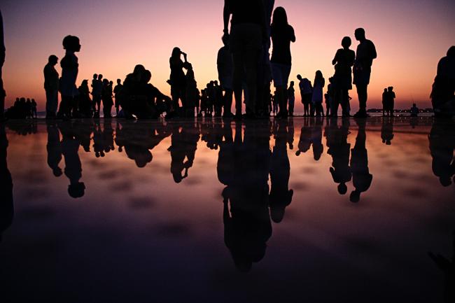 The silhouettes of many people are visible against a dim sunset. Their images are reflected on some body of water.