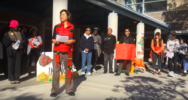 Rev. Deborah Lee, an Asian woman wearing a red shirt and red clerical shawl, speaks at a demonstration. 