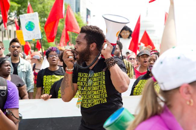A Black bearded man chants into a bullhorn at a demonstration. He is wearing a #BlackLivesMatter shirt.
